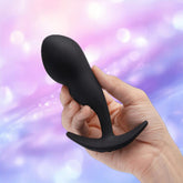 A hand holds the B-Vibe Vibrating P-Spot Snug Plug RC Large, a sleek black remote control anal plug by B-Vibe, against a soft, blurred background with pink, purple, and blue tones.